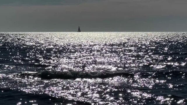 Panoramic view of ocean waves with sparkling sun reflection and sailboat on horizon.
