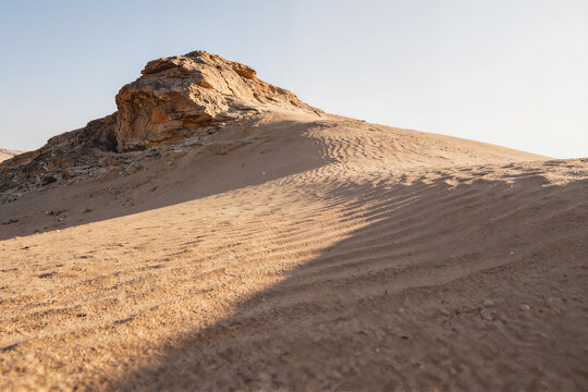 Vast desert with a rugged stratified rock outcrop rising from a sandy dune, wind-carved ripples and parallel grooves traversing the sand in warm tawny and rust hues