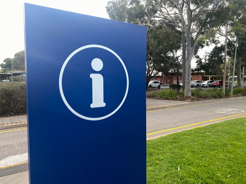 Blue sign showing white information symbol in a circle, offering guidance. Road, grass, trees, and cars in background