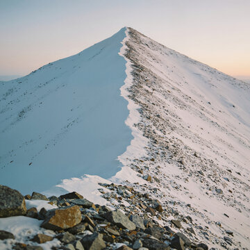 Sharp alpine ridge cutting diagonally against a gradient sky, a central line of jagged exposed rock and scree dividing smooth snowfields tinted blue and soft pink