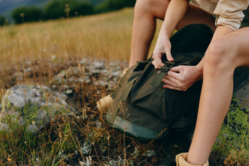 Obraz premium Young woman with short hair sits on grass in a natural field while opening a green backpack, enjoying a peaceful outdoor moment during a casual lifestyle hike.