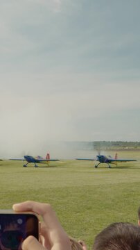 Vertical Screen: Crowds watch as aerobatic planes take off and perform tricks in the sky. Smoke trails fill the air as the show goes on at the airport during the daytime.