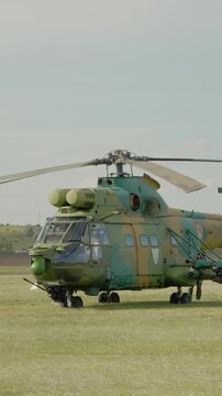 Vertical Screen: A Romanian combat helicopter sits on the grass at the airport and gets ready for takeoff during an air show. The day is clear, and other aircraft can be seen nearby.
