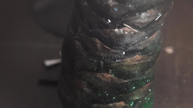 Liquid green epoxy resin with glitter filler slowly fills a transparent mold around a pine cone in workshop, macro vertical close-up.