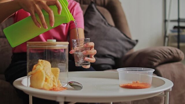 Close-up of woman pouring water into drinking glass on white table while snacking at home