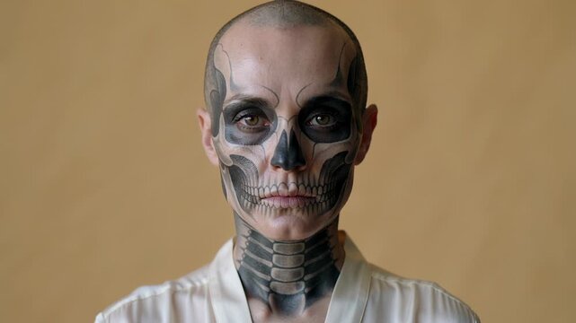 Turning shaved-head model shifting head, centering gaze at beige wall, in light robe, skull makeup