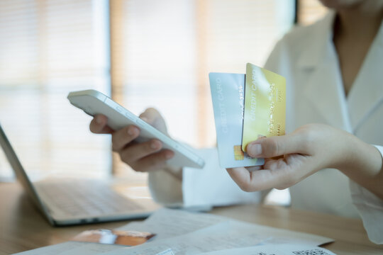 Person holds phone and credit card at desk. Light filters through blinds behind. Office setting suggests modern digital payments. Focus on hands implies financial transactions. Suitable for e-commerce