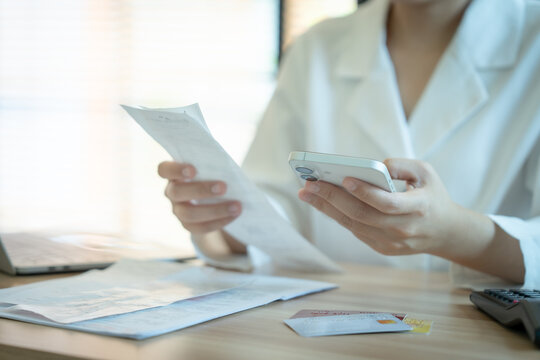 Woman in white coat holds phone and bill. She reviews charges while standing at desk. Soft light illuminates her focused expression. Documents and calculator sit beside her.
