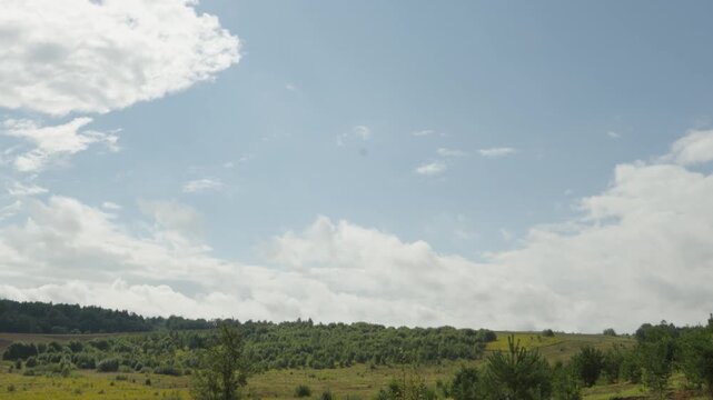 Fluffy white clouds drift slowly across a bright blue sky above a scenic rolling green countryside landscape with young trees and open fields.