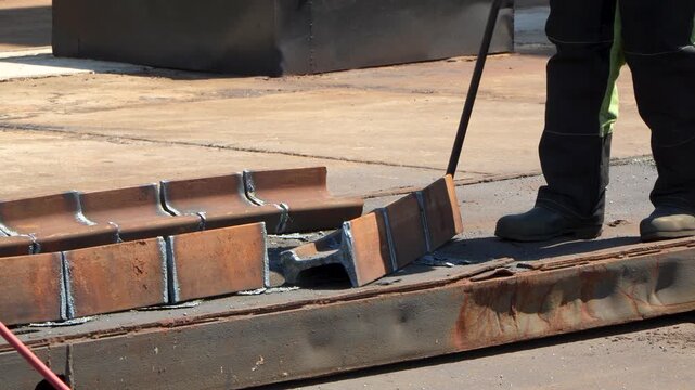 An industrial worker wearing protective gear and safety boots uses a hook tool to adjust and move heavy cut steel rail segments outdoors on a metal platform.