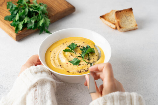 Woman eating creamy pumpkin soup with cream at a light kitchen table, hands in white knitted sweater, cozy autumn homemade comfort food