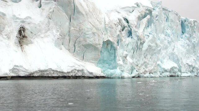 Glacier Front With Blue Ice Above Calm Gray Water. Wide cold-water view shows a steep fractured glacier face with pale blue crevasses and white snow, meeting a smooth dark marine surface.