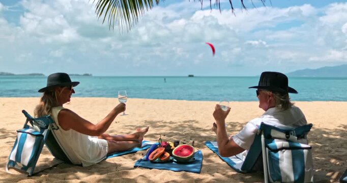 Senior couple enjoying a tropical beach vacation. Sitting in portable chairs under palm shade. Drinking wine and having a picnic with fresh fruit on the sand by the turquoise ocean in Samui, Thailand
