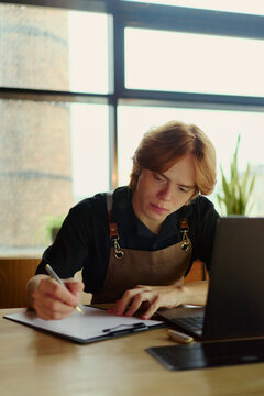Vertical image of Caucasian young small business owner writing notes while working on laptop. Ideal for small business management, entrepreneurship, and cafe accounting concepts