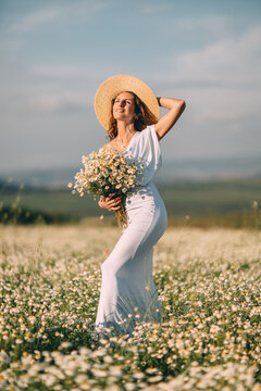 Sunlit meadow portrait featuring young woman in white dress holding bouquet of Matricaria chamomilla flowers during warm daytime