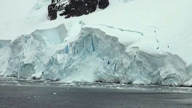 Snowy Glacier Cliff With Blue Ice and Dark Rock Above. A tall frozen coastal face rises from dark water, combining fractured blue-white ice, snow fields and exposed black rock along the upper slope.