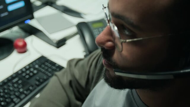 Young biracial man with glasses wearing headset talking to client and typing on computer keyboard. Useful for customer service training, technical support hotline, and telemarketing ads