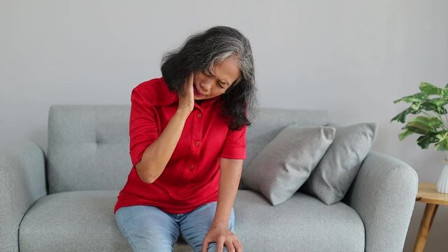 Senior Asian woman sitting on a sofa, touching her cheek with a painful expression, illustrating toothache, dental pain, jaw discomfort, and oral health problems.