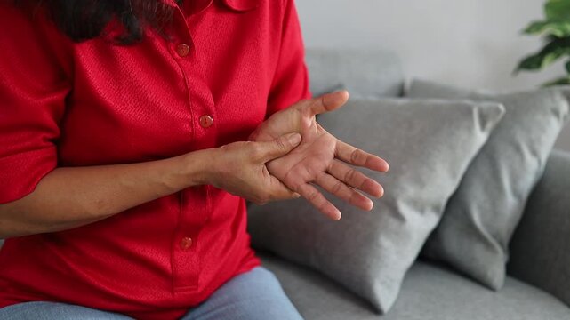 Senior Asian woman experiencing hand pain and massaging her palm due to numbness and joint discomfort at home.