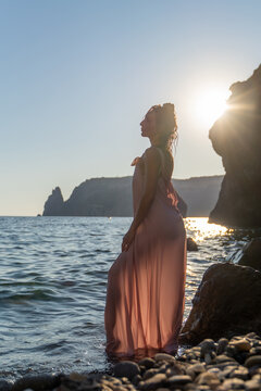 Woman in pink dress posing gracefully by rocky sea shore during sunset light
