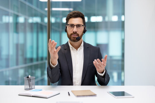A young businessman wearing a headset conducts an online video meeting; he sits at an office desk and explains while gesturing with his hands toward the camera