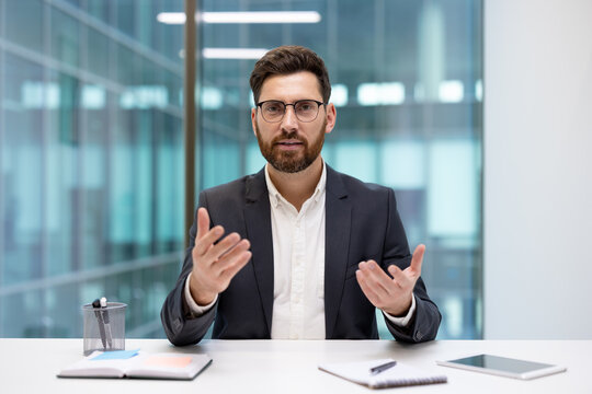 A young businessman is conducting an online video meeting; he is sitting at an office desk and explaining something while gesturing with his hands toward the camera
