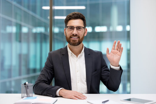 A portrait of a young, smiling man in a suit and glasses, sitting at an office desk and waving his left hand at the camera