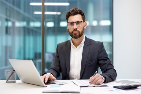 A portrait of a focused young businessman in a suit and glasses, working at an office desk with a laptop and documents, looking at the camera