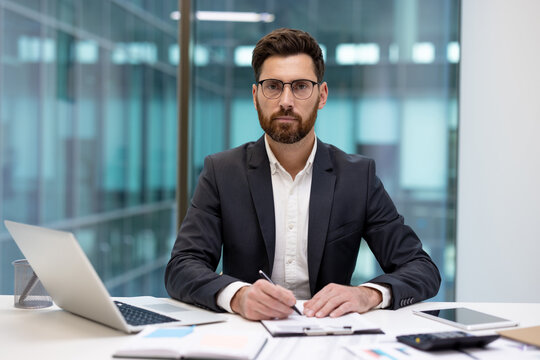 A portrait of a serious young businessman in a suit and glasses, sitting at an office desk with a laptop and documents, holding a pen and looking at the camera