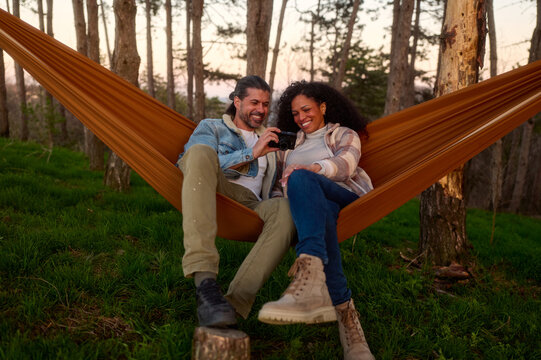 Diverse couple relaxing in a hammock in a sunlit forest, smiling as they share and review photos on a compact digital camera during a peaceful outdoor getaway