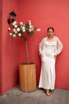 Young woman in white satin dress stands gracefully against a vibrant coral red wall outdoors