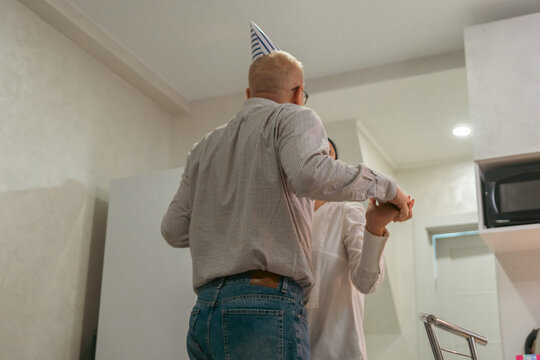 Here's a title attempting to follow all instructions: Birthday Party Couple Dancing Home: Man wearing party hat joyfully dances with woman in home kitchen celebrating birthday.