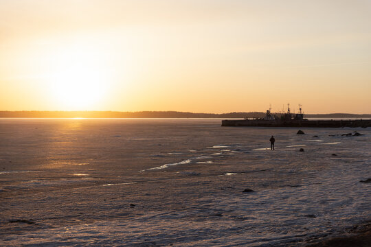A solitary figure walks across a frozen sea near a harbor as the low winter sun casts warm golden light and long shadows over icy shoreline and distant boats at dusk