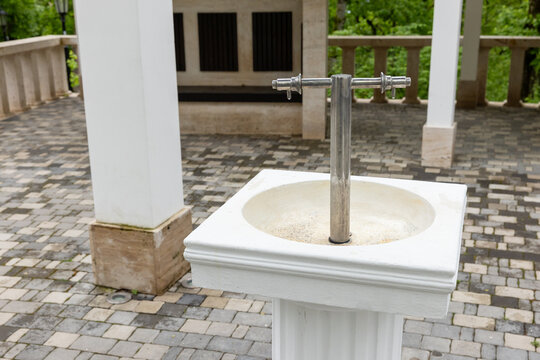 Outdoor drinking fountain featuring a stainless steel spout mounted on a white pedestal sink under a covered pavilion, set on stone pavers with green foliage visible in the background