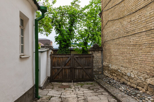 An old stone courtyard framed by a weathered wooden gate, a brick wall and a white plaster house with a green downspout, opening to leafy trees beyond. Rustic, quiet urban charm. Pyatigorsk, Russia