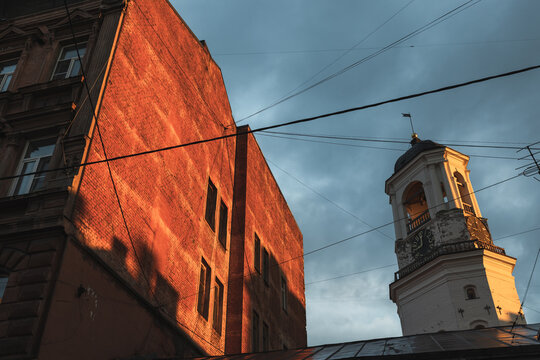 Historic clock tower of Vyborg old town rises beside a red brick building as warm golden-hour sunlight illuminates the facade, contrasting with moody blue clouds