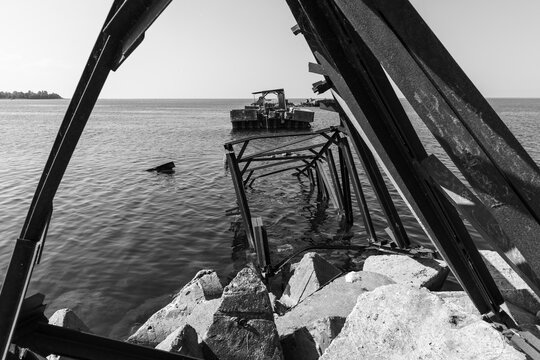 Perspective view of a rusted metal structure extends into coastal waters beside a rocky shoreline, evoking industrial decay and rugged seaside atmosphere. Broken bridge of Battery bay, Russia