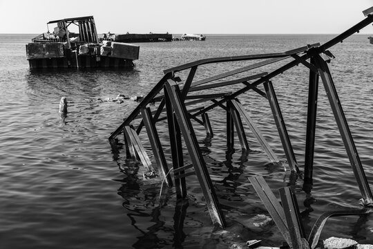 Black and white photo of a rusted metal structure extends into coastal waters beside a rocky shoreline, evoking industrial decay and rugged seaside atmosphere. Broken bridge of Battery bay, Russia