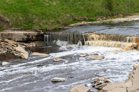 A scenic small waterfall cascades over layered rocks into a shallow, fast-flowing river. Sablinsky Waterfall on the Sablinka River in the Tosnensky District of the Leningrad Region, Russia