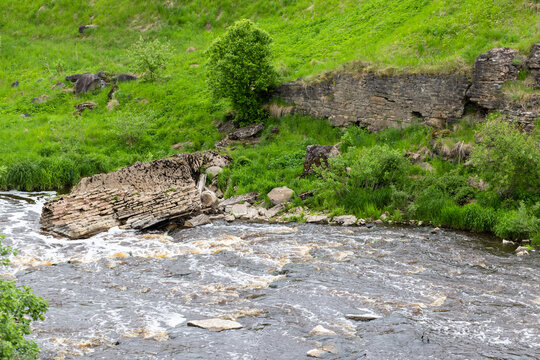 Ruined stone wall, bridge and collapsed masonry sit at the edge of a fast-flowing river, surrounded by lush green grass, bushes and rocks. Sablinka River, Russia