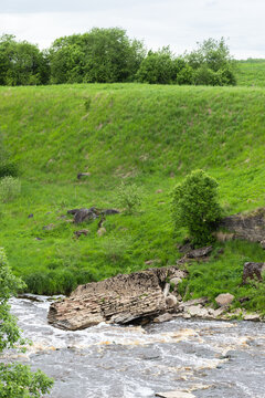 Vertical photo displaying ruined stone wall, bridge and collapsed masonry sit at the edge of a fast-flowing river, surrounded by lush green grass, bushes and rocks. Sablinka River, Russia