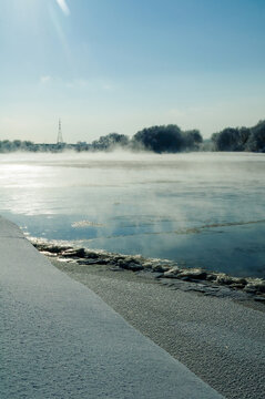 Winter landscape background. A wide river covered with mist and floating ice. Wet pebble shore in the foreground and frosted trees on the distant bank