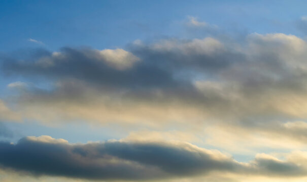 Panoramic sunset sky background. Layered dark grey clouds with warm golden light on the lower edges. A blue sky visible between the soft formations above the horizon