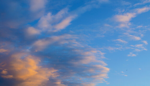 Dramatic sunset sky panoramic background. Warm golden lit clouds on the left side of a blue sky. Soft purple tones and delicate cloud layers stretching across the evening atmosphere
