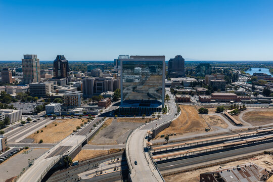 Aerial view shows the downtown Sacramento skyline, featuring modern commercial buildings, railway tracks, highway overpasses, and the Sacramento River in California.