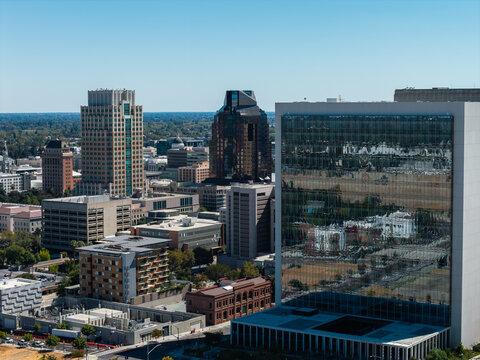 Aerial perspective showcases the urban skyline of downtown Sacramento, California, displaying office towers, glass facades, and the distant California State Capitol dome on a clear sunny day.