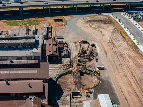 Aerial view shows the historic railway turntable and maintenance workshops at the Sacramento Railyards in California, alongside an adjacent highway and dirt lot.