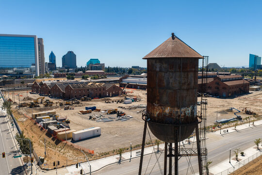 Aerial perspective of the historic Sacramento Railyards redevelopment site with a weathered water tower, renovated brick shops, and the downtown city skyline visible under clear daytime skies.