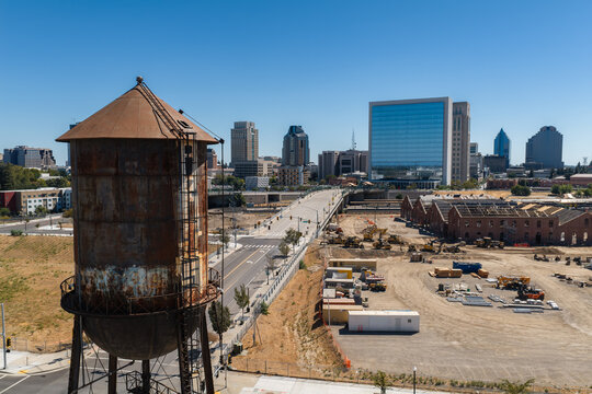 Aerial view of the historic rusted water tower and urban redevelopment site at the Sacramento Railyards in downtown Sacramento, California, with city skyline under clear sky.