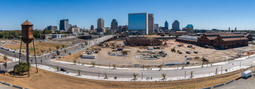 Aerial panorama captures construction and redevelopment across the historic Sacramento Railyards with the downtown skyline under a clear daytime sky in California.